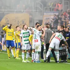 Piłkarze w białych i żółtych strojach piją wodę na boisku, w tle kibice.