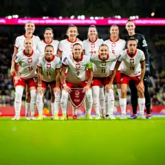 Piłkarki Polski gotowe do meczu z Rumunią. Drużyna w białych strojach z orłem na piersiach pozuje na tle trybun.