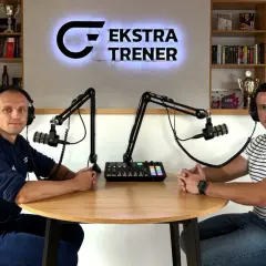 Dwóch mężczyzn w słuchawkach siedzi przy stole, gotowych do nagrania podcastu "Ekstra Trener". W tle półki z książkami i trofeami, tworząc atmosferę klubu kokosa.