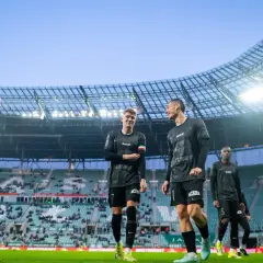 Piłkarze w czarnych strojach na murawie stadionu. Jeden z nich pyta: "walkower co to?", ale koledzy skupiają się na grze.