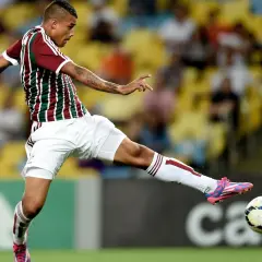 Piłkarz w koszulce Fluminense kopie piłkę na tle trybun.