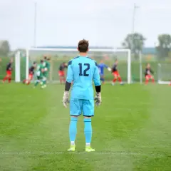 Golkiper w błękitnym stroju z numerem 12 czeka na akcję na boisku. W tle inni zawodnicy i bramka.