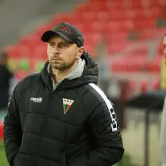 Trener GKS Tychy w czapce z napisem "Tychy" i kurtce z herbem klubu. W tle trybuny stadionu.