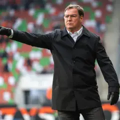 Trener w czarnej kurtce i rękawiczkach wskazuje palcem, jak doświadczony selekcjoner kieruje grą.