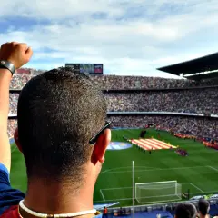 Widok z trybun stadionu na murawę, gdzie kibice tworzą flagę. W powietrzu unosi się hymn Barcelony, a kibic z podniesioną pięścią czeka na mecz.