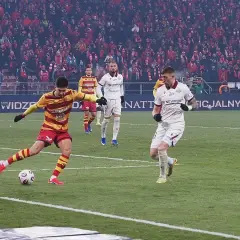 Piłkarz w żółto-czerwonych barwach strzela gola, podczas gdy zawodnik Jagiellonii Białystok w białej koszulce biegnie obok. Meczu Widzew Łódź – Jagiellonia Białystok.