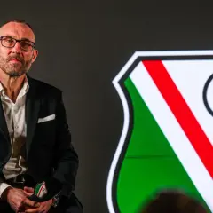 Mężczyzna w garniturze, nowy trener Legii Warszawa, siedzi obok herbu klubu.