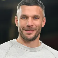 Uśmiechnięty Lukas Podolski w szarej bluzie. Jego majątek budowany latami kariery piłkarskiej i biznesów.