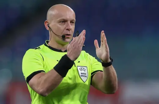 Sędzia piłkarski w jaskrawej koszulce z emblematem UEFA, gestykuluje. Ciekawe, ile zarabia sędzia piłkarski za takie emocje na boisku.