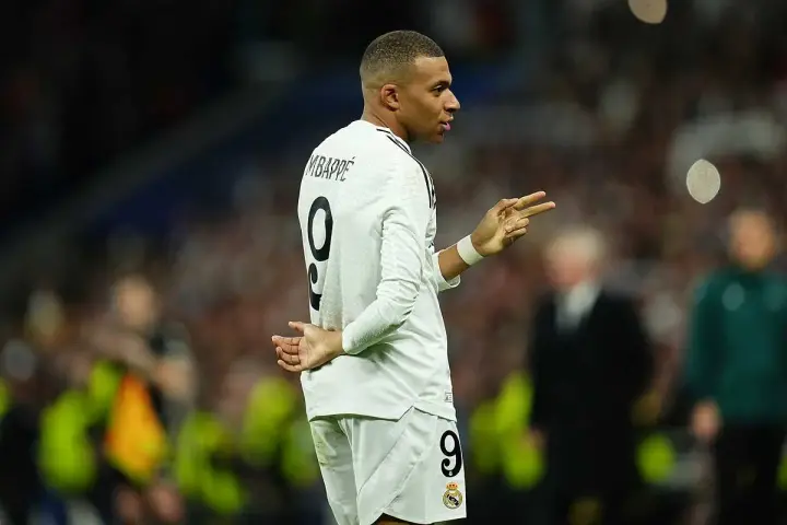Kylian Mbappé, młody piłkarz, w białej koszulce z numerem 9, gestykuluje na boisku. Jego wiek to zaledwie 24 lata.