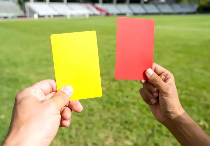 Dwie ręce trzymają żółtą i czerwoną kartkę na tle zielonej murawy stadionu. Decyzja sędziego zależy od kolorów kart.