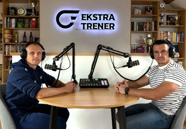 Dwóch mężczyzn w słuchawkach siedzi przy stole, gotowych do nagrania podcastu "Ekstra Trener". W tle półki z książkami i trofeami, tworząc atmosferę klubu kokosa.