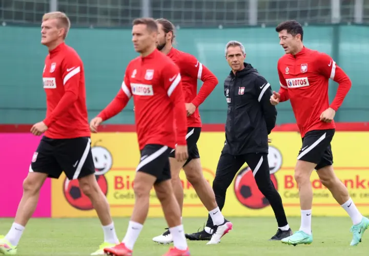 Piłkarze reprezentacji Polski w czerwonych strojach i trener w czarnej kurtce podczas treningu. Lewandowski i inni zawodnicy w akcji.