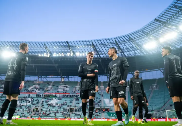 Piłkarze w czarnych strojach na murawie stadionu. Jeden z nich pyta: "walkower co to?", ale koledzy skupiają się na grze.