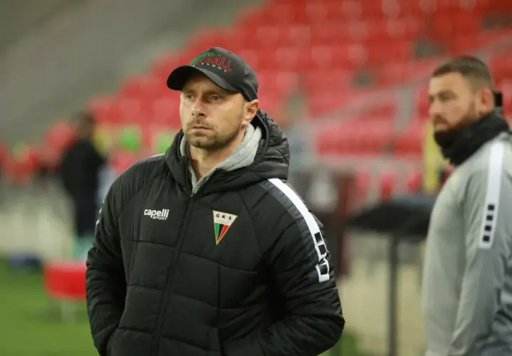 Trener GKS Tychy w czapce z napisem "Tychy" i kurtce z herbem klubu. W tle trybuny stadionu.
