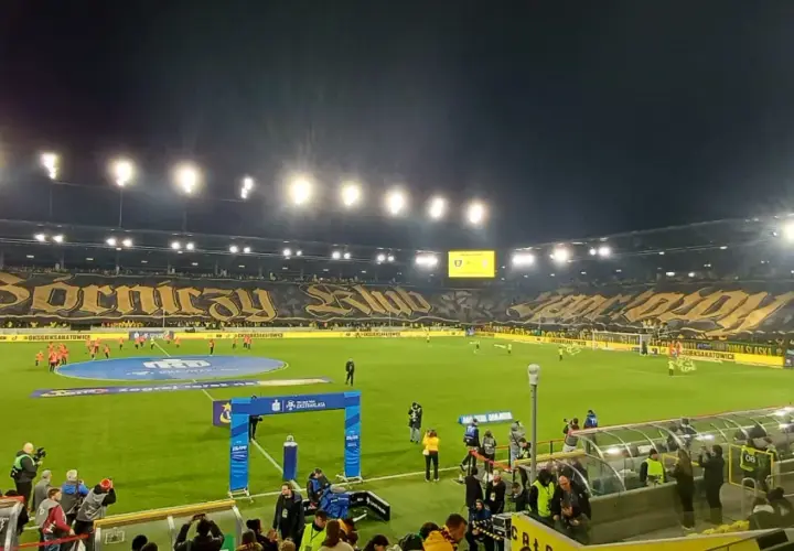 Stadion GKS Katowice podczas meczu z Górnikiem Zabrze. Trybuny wypełnione kibicami, wielka oprawa z napisem "Górniczy Klub".