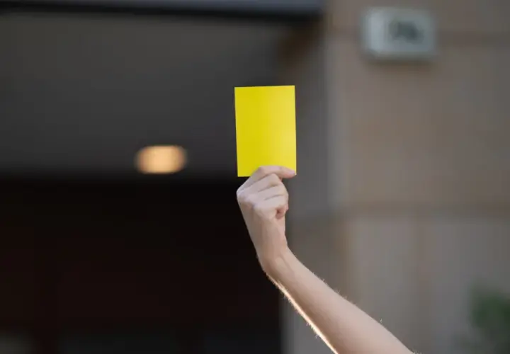 Ręka unosi żółtą kartkę. Może to symbol ostrzeżenia lub przypomnienia.