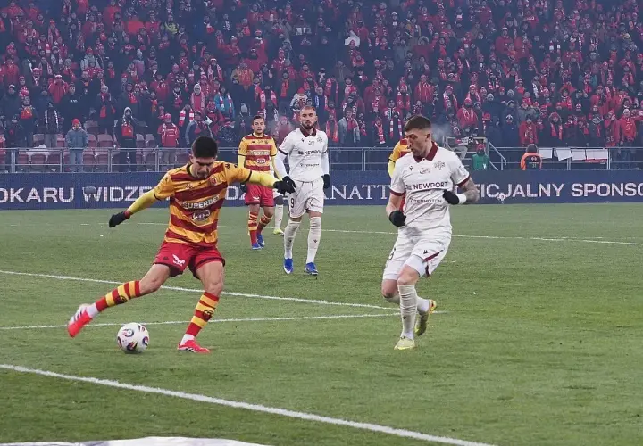 Piłkarz w żółto-czerwonych barwach strzela gola, podczas gdy zawodnik Jagiellonii Białystok w białej koszulce biegnie obok. Meczu Widzew Łódź – Jagiellonia Białystok.