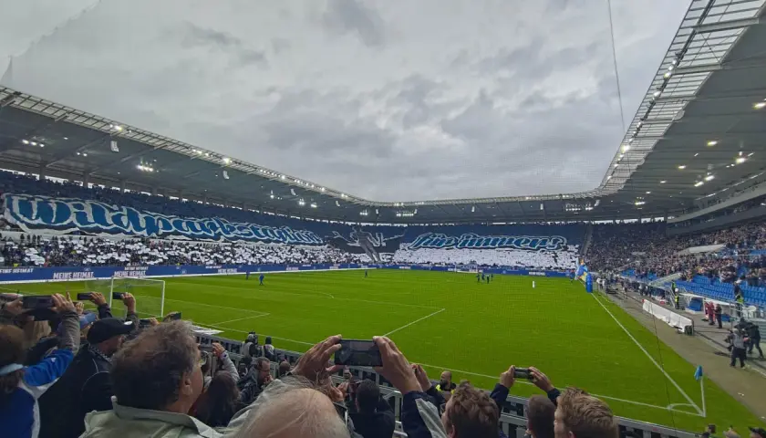 Trybuny stadionu wypełnione kibicami Karlsruher SC. Fani tworzą ogromną oprawę z napisami i sylwetką zawodnika.