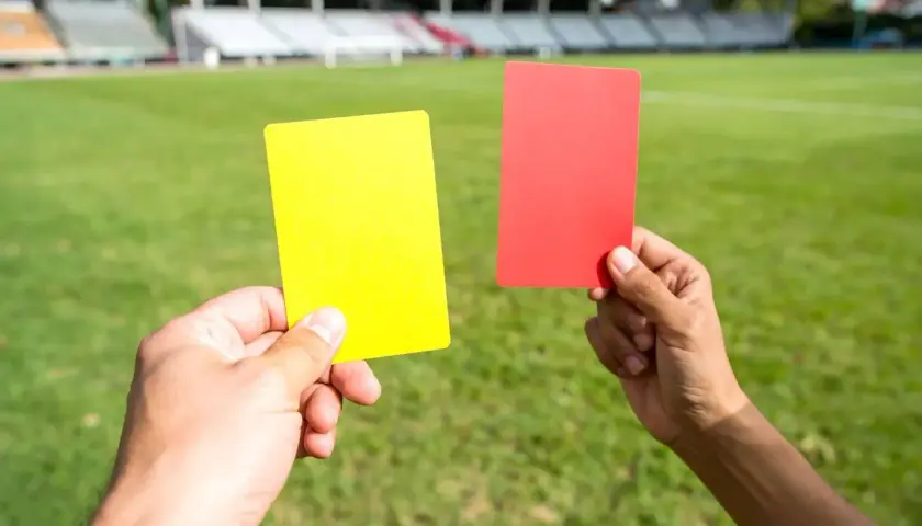 Dwie ręce trzymają żółtą i czerwoną kartkę na tle zielonej murawy stadionu. Decyzja sędziego zależy od kolorów kart.