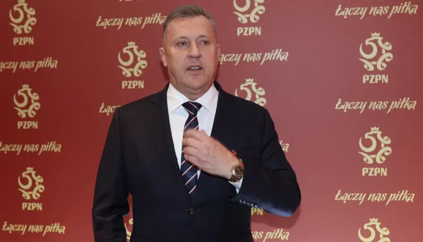 Prezes PZPN w garniturze, poprawiający krawat. Tło ozdobione logotypami PZPN i hasłem "Łączy nas piłka".