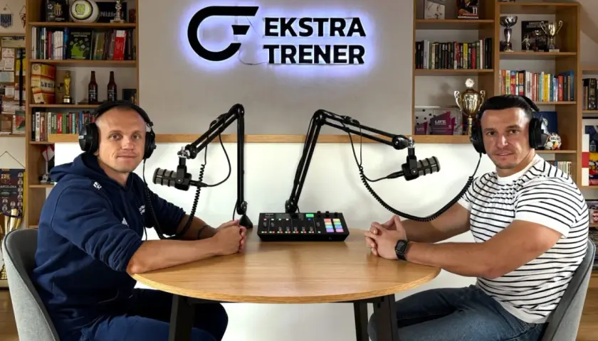 Dwóch mężczyzn w słuchawkach siedzi przy stole, gotowych do nagrania podcastu "Ekstra Trener". W tle półki z książkami i trofeami, tworząc atmosferę klubu kokosa.