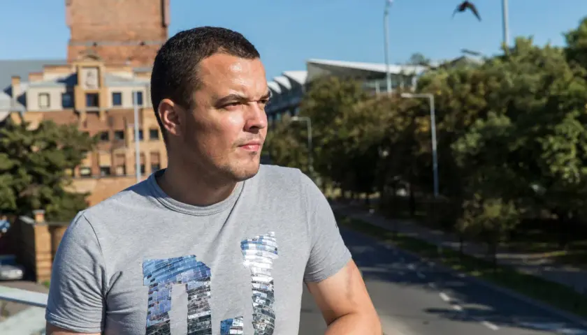 Dawid Janczyk w szarej koszulce z błyszczącym napisem, na tle miejskiego krajobrazu z budynkami i drzewami.