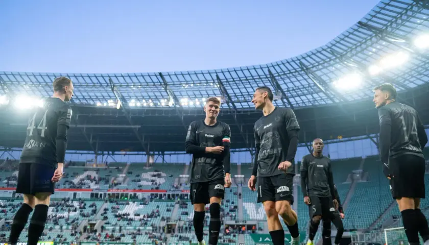 Piłkarze w czarnych strojach na murawie stadionu. Jeden z nich pyta: "walkower co to?", ale koledzy skupiają się na grze.