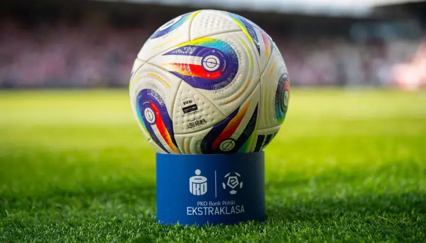 Piłka do piłki nożnej na niebieskim postumencie z logo Ekstraklasy na tle stadionu.