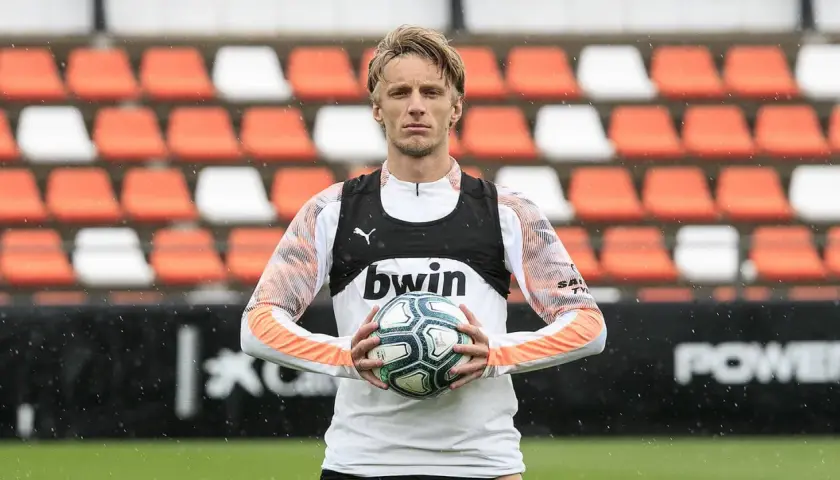 Piłkarz z blond włosami trzyma piłkę, ubrany w białą koszulkę z pomarańczowymi rękawami i czarną kamizelkę z logo bwin. W tle pomarańczowo-białe trybuny.