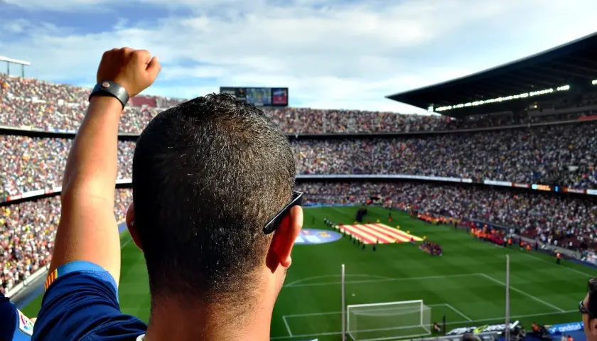 Widok z trybun stadionu na murawę, gdzie kibice tworzą flagę. W powietrzu unosi się hymn Barcelony, a kibic z podniesioną pięścią czeka na mecz.