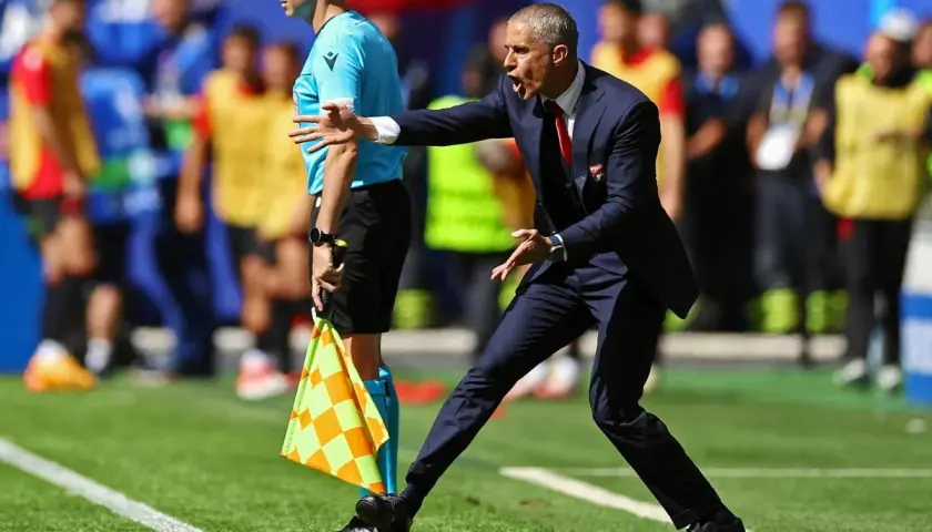 Sylvinho, trener w garniturze, gestykuluje na boisku. Obok stoi sędzia liniowy z flagą.