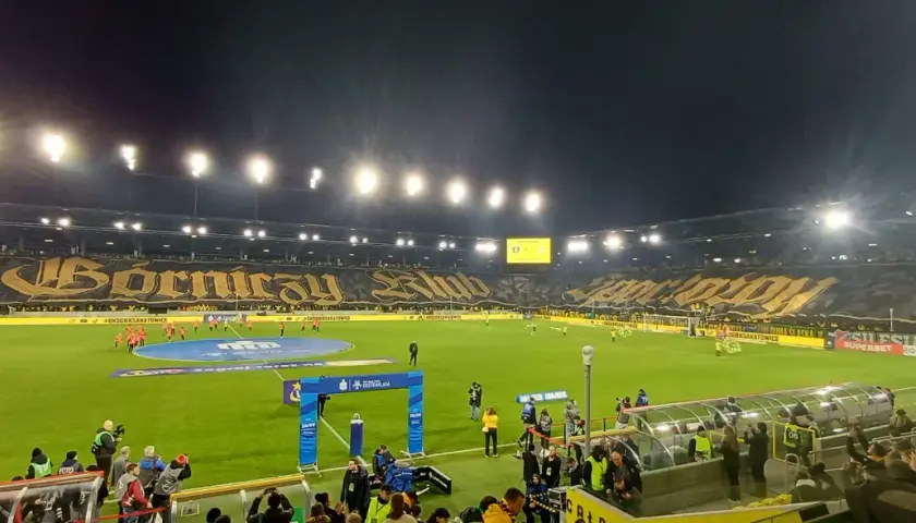 Stadion GKS Katowice podczas meczu z Górnikiem Zabrze. Trybuny wypełnione kibicami, wielka oprawa z napisem "Górniczy Klub".