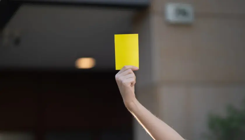 Ręka unosi żółtą kartkę. Może to symbol ostrzeżenia lub przypomnienia.