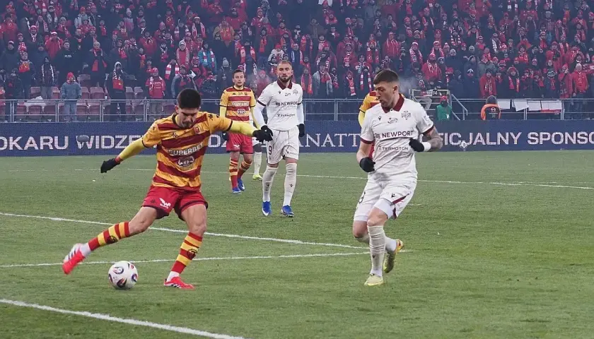 Piłkarz w żółto-czerwonych barwach strzela gola, podczas gdy zawodnik Jagiellonii Białystok w białej koszulce biegnie obok. Meczu Widzew Łódź – Jagiellonia Białystok.