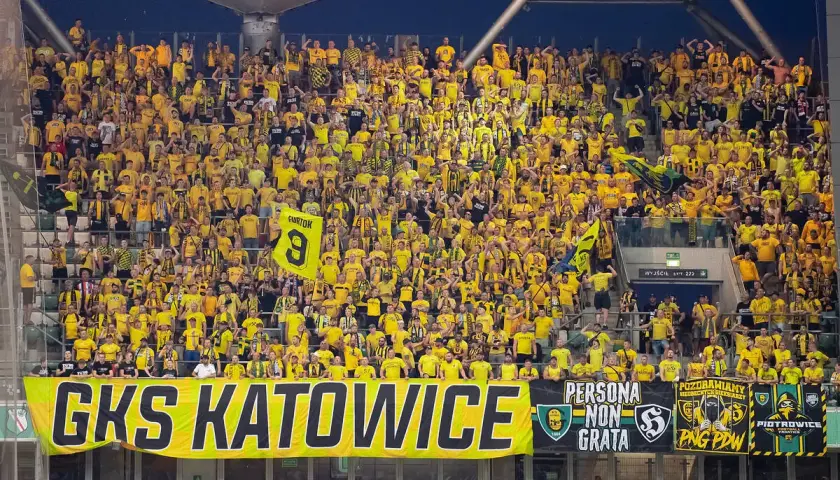 Tłum kibiców GKS Katowice w żółtych barwach, z transparentem "GKS KATOWICE" i flagami.