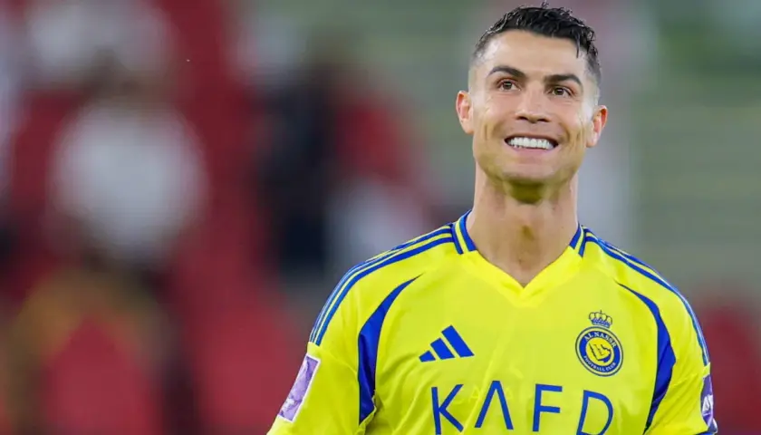 Uśmiechnięty Cristiano Ronaldo w żółtej koszulce z niebieskimi pasami, grający w klubie Al Nassr.