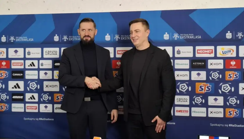 Dwóch mężczyzn w garniturach ściska sobie dłonie na tle ścianki sponsorskiej Rakowa Częstochowa. Zapowiada to nowe, ekscytujące transfery!