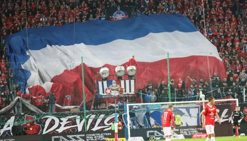 Oprawa meczu Ruch Chorzów – Wisła Kraków. Ogromna flaga w barwach biało-czerwono-niebieskich zakrywa trybuny, a pod nią bębny i kibice.