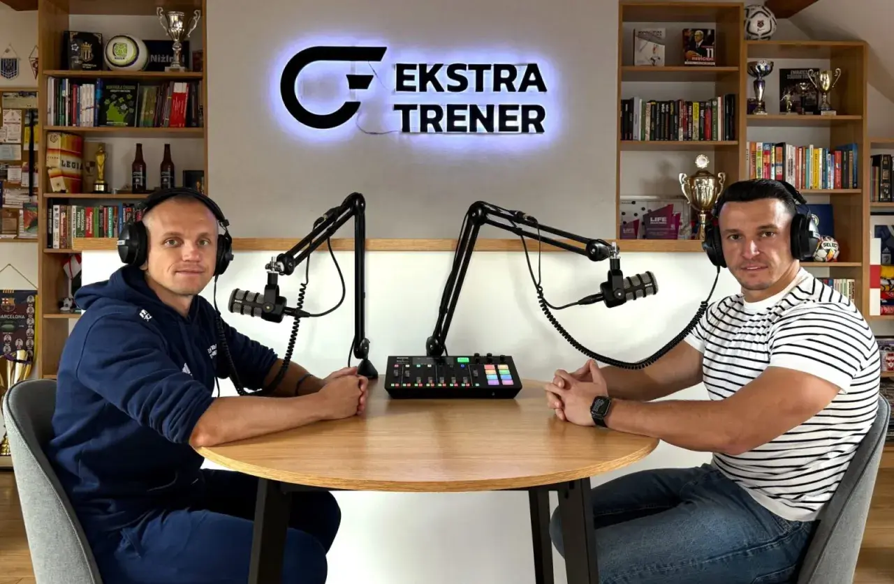 Dwóch mężczyzn w słuchawkach siedzi przy stole, gotowych do nagrania podcastu "Ekstra Trener". W tle półki z książkami i trofeami, tworząc atmosferę klubu kokosa.