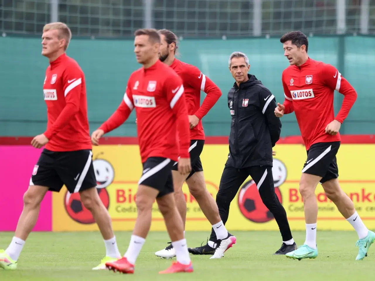 Piłkarze reprezentacji Polski w czerwonych strojach i trener w czarnej kurtce podczas treningu. Lewandowski i inni zawodnicy w akcji.