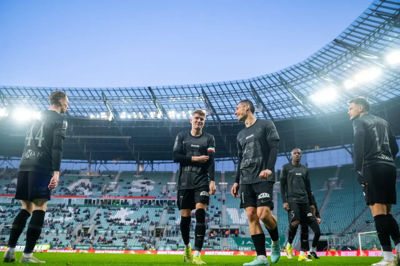 Piłkarze w czarnych strojach na murawie stadionu. Jeden z nich pyta: "walkower co to?", ale koledzy skupiają się na grze.