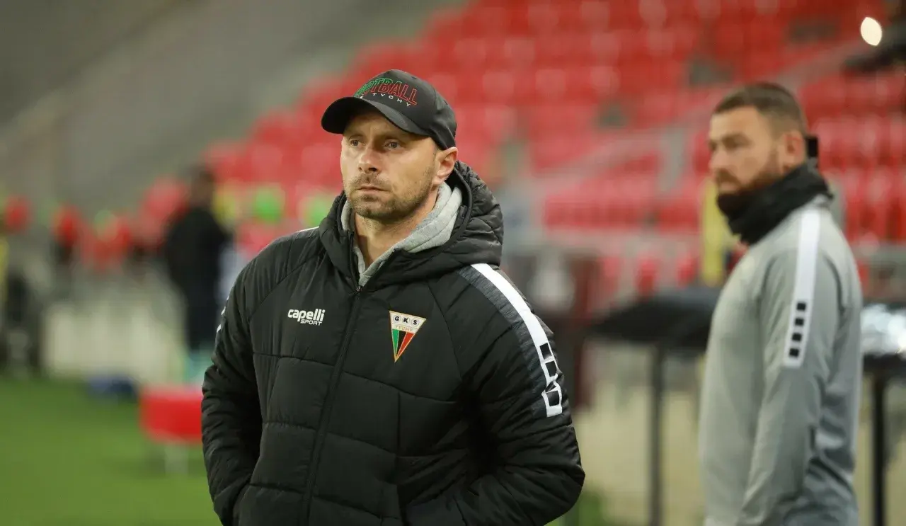 Trener GKS Tychy w czapce z napisem "Tychy" i kurtce z herbem klubu. W tle trybuny stadionu.