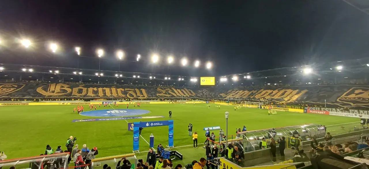 Stadion GKS Katowice podczas meczu z Górnikiem Zabrze. Trybuny wypełnione kibicami, wielka oprawa z napisem "Górniczy Klub".