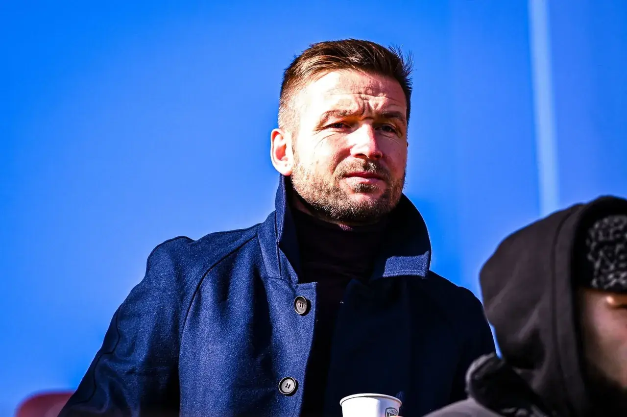 Piast Gliwice trener w granatowym płaszczu, zamyślony pod błękitnym niebem.
