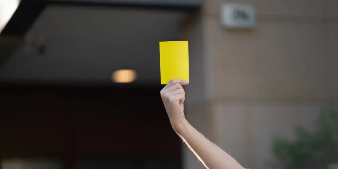 Ręka unosi żółtą kartkę. Może to symbol ostrzeżenia lub przypomnienia.