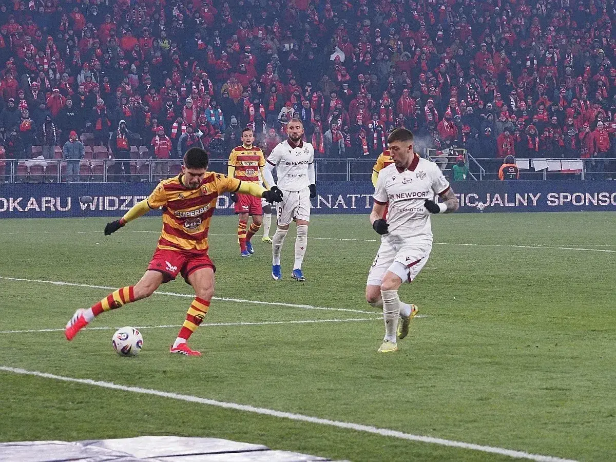 Piłkarz w żółto-czerwonych barwach strzela gola, podczas gdy zawodnik Jagiellonii Białystok w białej koszulce biegnie obok. Meczu Widzew Łódź – Jagiellonia Białystok.