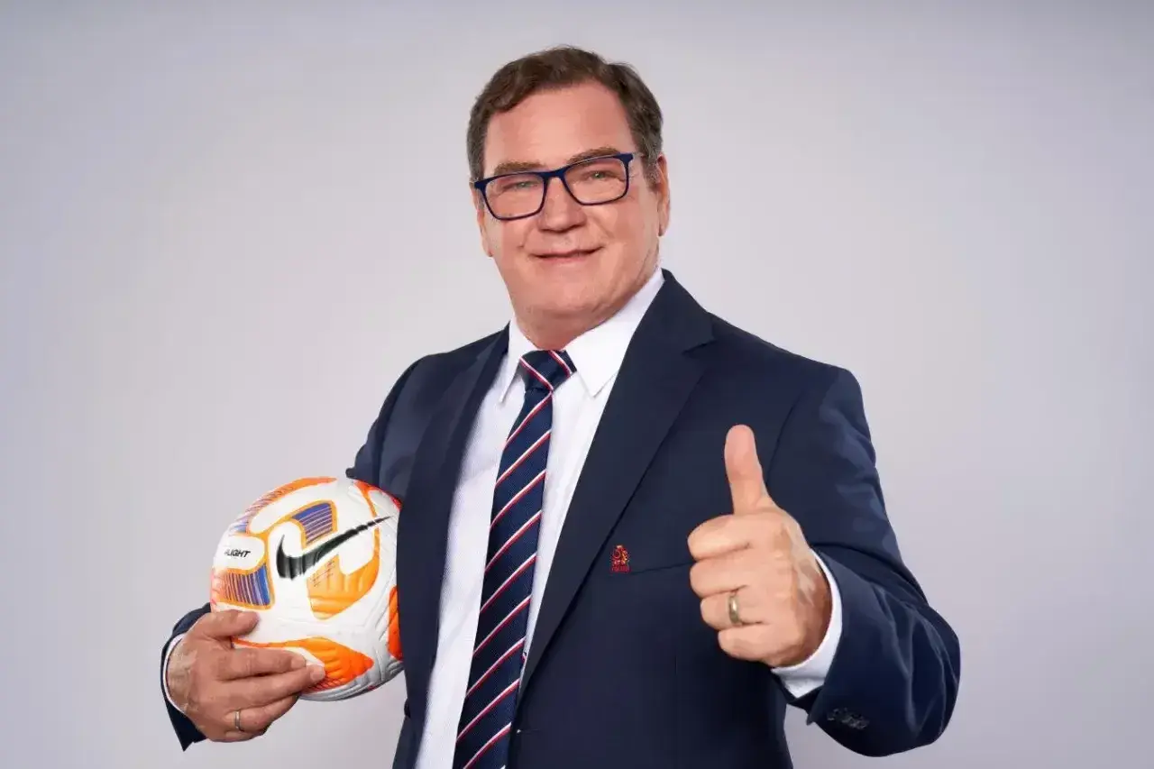 Uśmiechnięty trener reprezentacji Polski w garniturze trzyma piłkę i pokazuje kciuk w górę.