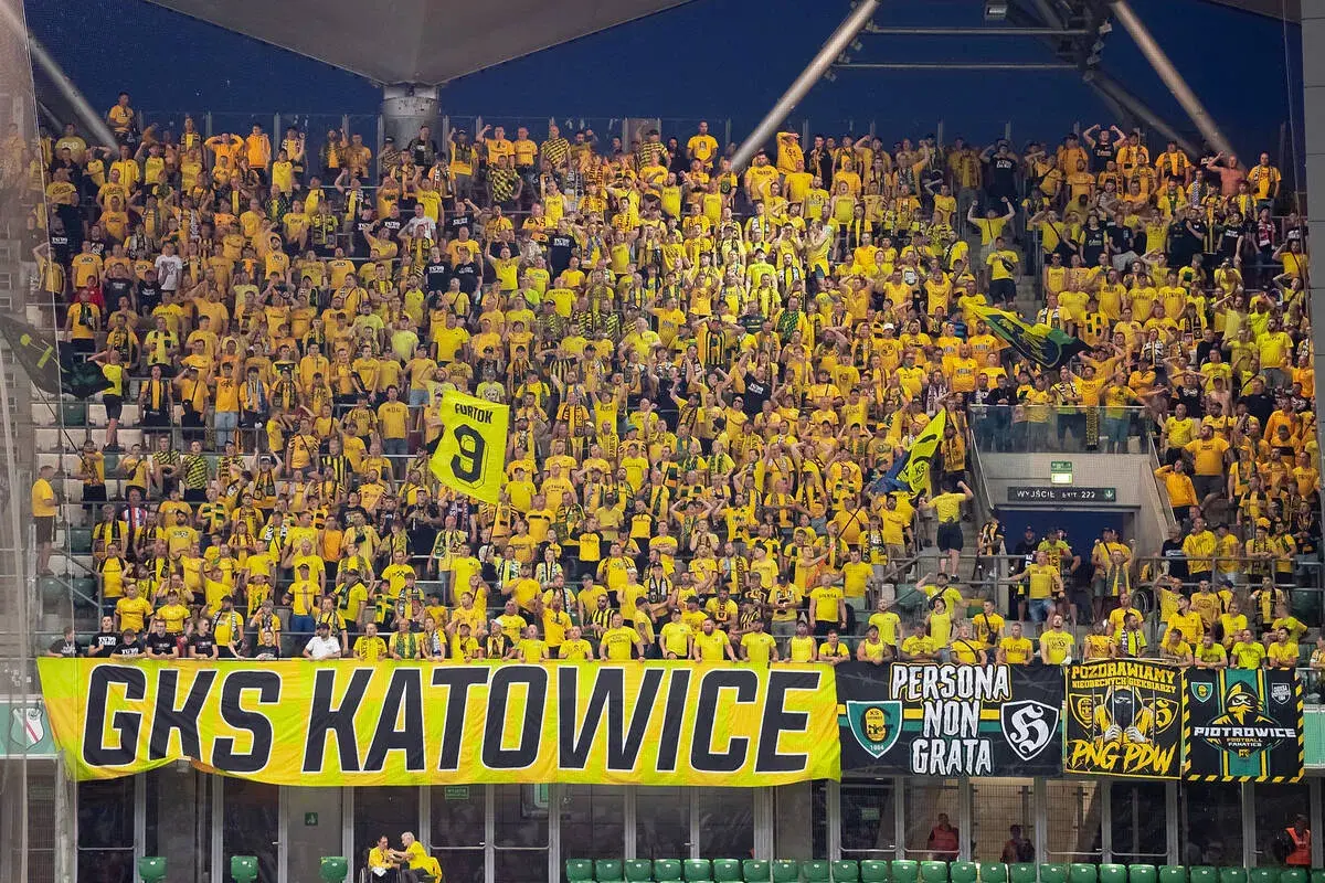 Tłum kibiców GKS Katowice w żółtych barwach, z transparentem "GKS KATOWICE" i flagami.