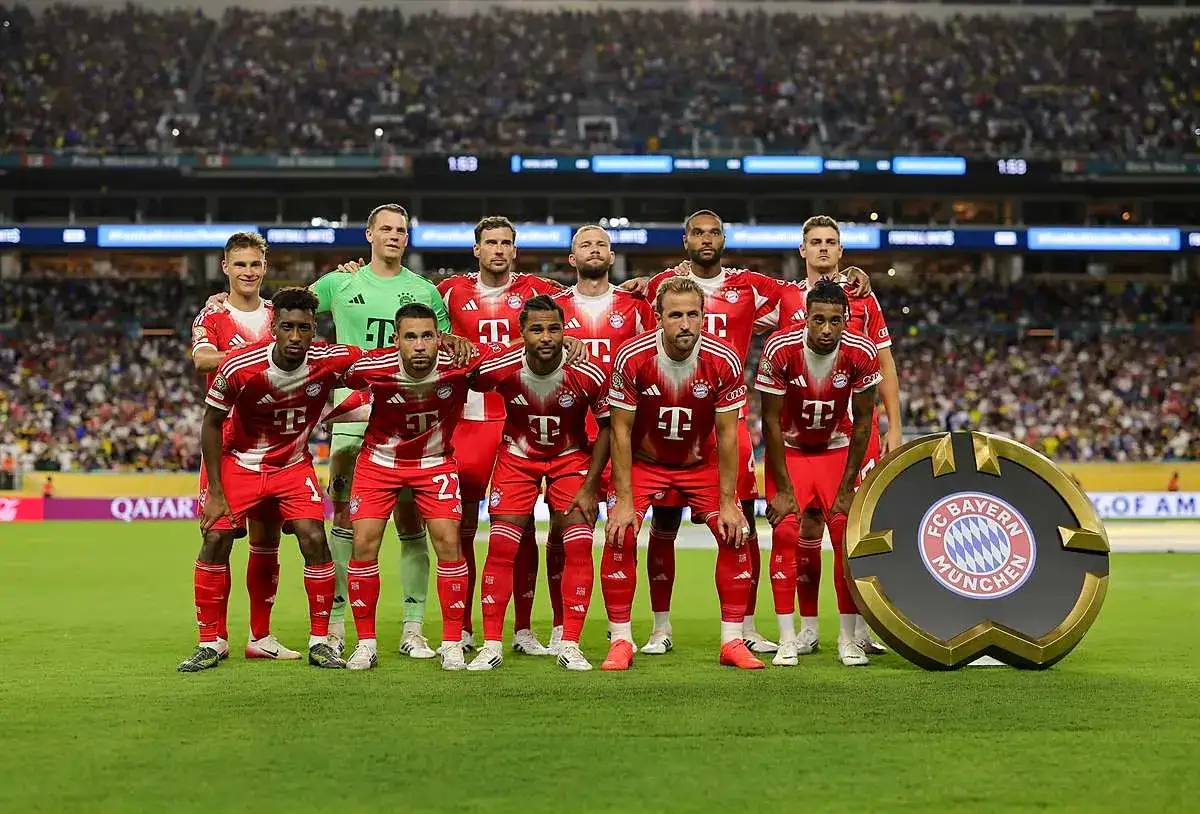 Drużyna Bayern Monachium – zawodnicy w czerwonych strojach pozują do zdjęcia na tle trybun pełnych kibiców. Obok nich stoi okrągłe logo klubu.